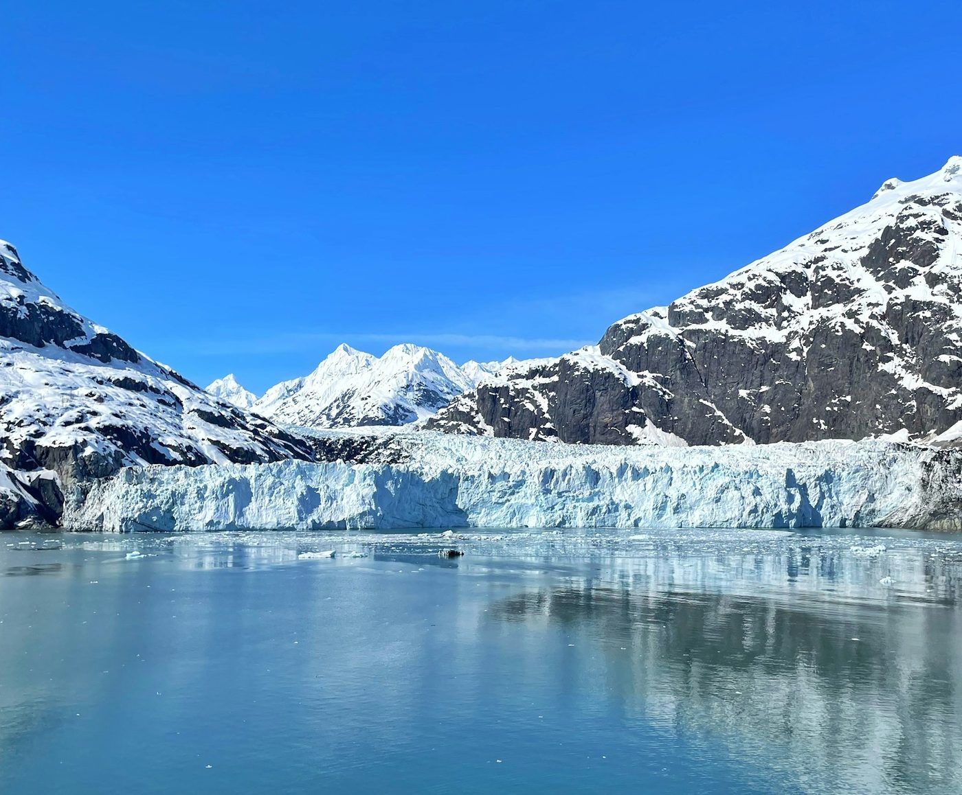 Tidewater glacier front with snow-covered Alaskan mountains under a clear blue sky, seen from the water