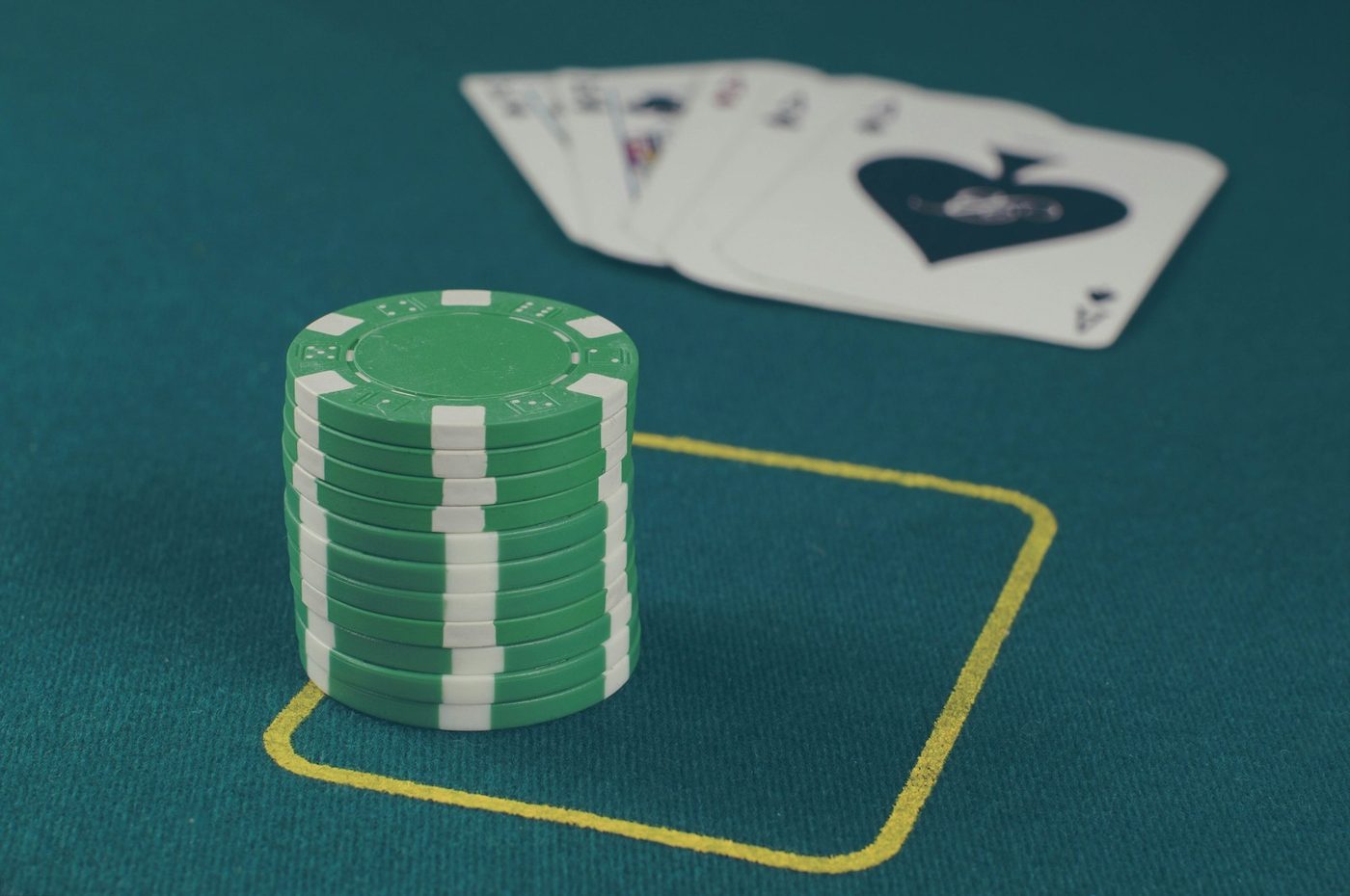 Stack of green casino chips inside a yellow betting box on green felt with playing cards, ace of spades showing