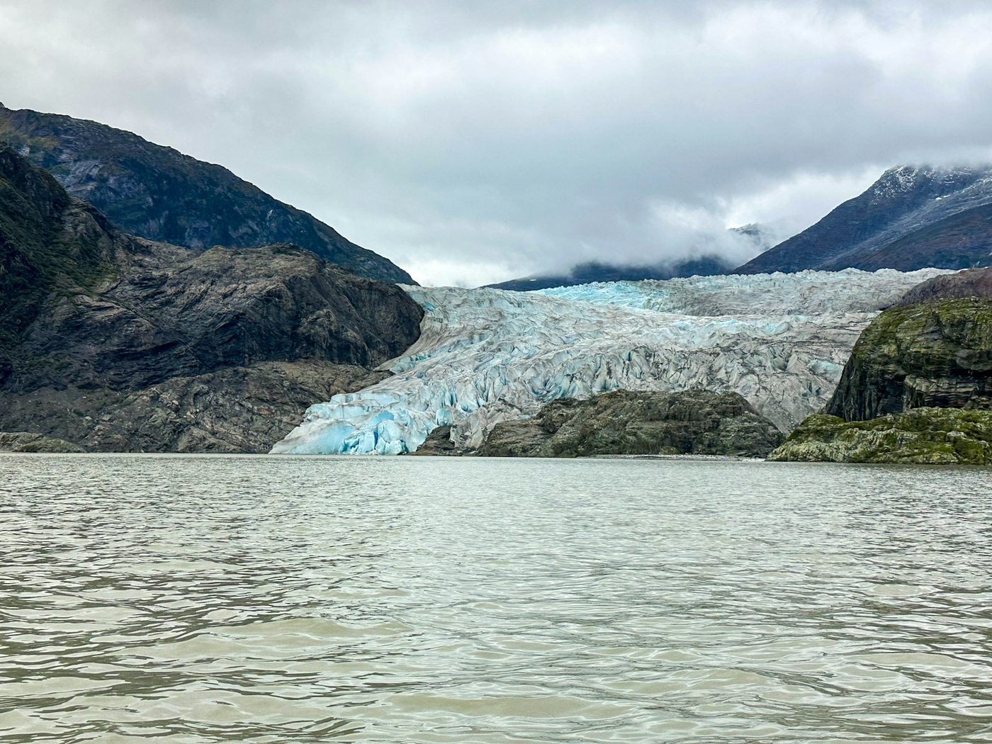 Alaskan glacier flowing between dark mountains under low clouds, photographed from water level as if from a tender boat