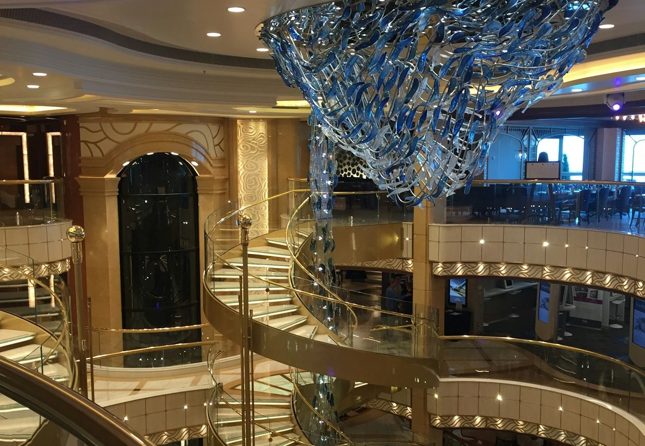 Multi-deck cruise ship atrium with gold spiral staircase and glass sculpture — the suite-class ambience cruise lines sell at double fare